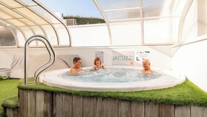 A hot tub with three people, surrounded by a wooden deck and artificial grass, under a glass canopy. - Carva - Confort 1 TV - 2p (La Guérinière)