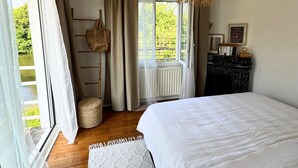 Een slaapkamer met een groot bed, een raam met gordijnen, een radiator, een houten vloer en een boekenplank.