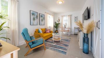 Uma sala de estar com um sofá amarelo, uma poltrona azul, uma mesa de centro e uma TV fixada na parede.