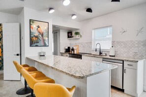 A modern kitchen with a central island, white cabinetry, and a large window with blinds. - Birds of Paradise: Relaxing Coastal Escape (New Smyrna Beach)