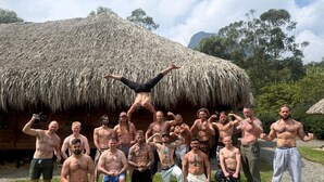 Een groep mannen zonder shirt poseert voor een hut met rieten dak, met een grasveld en bomen op de achtergrond.