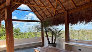 A wooden table and chairs under a thatched roof with string lights, overlooking a lush landscape. - Pool Rooftop Gym Tulum 2BR Penthouse A5 (Tulum)