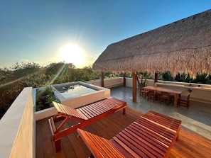 Eine Dachterrasse mit Pool, Holzliegen, einem Esstisch und einem Reetdach.