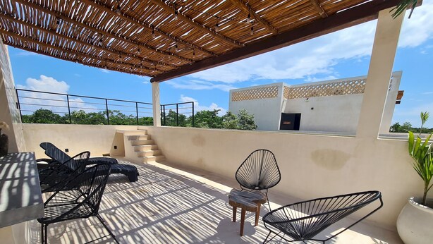 Un área de estar al aire libre con techo, que incluye una mesa, sillas, una escalera y una vista de un edificio al fondo.
