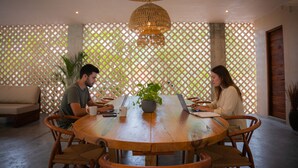A round wooden table with two people working on laptops, surrounded by wicker chairs, in a room with a woven ceiling light fixture and a lattice wall. - Jungle Escape Studio Quiet Cozy Gym Pool C5 (Tulum)
