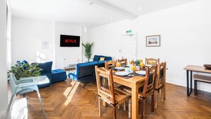Una sala de estar moderna con comedor, un sofá azul, sillas de madera y un televisor que muestra 'NETFLIX'.