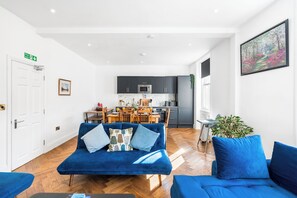 Een moderne woonkamer met een blauwe bank, een eethoek en een keuken op de achtergrond.