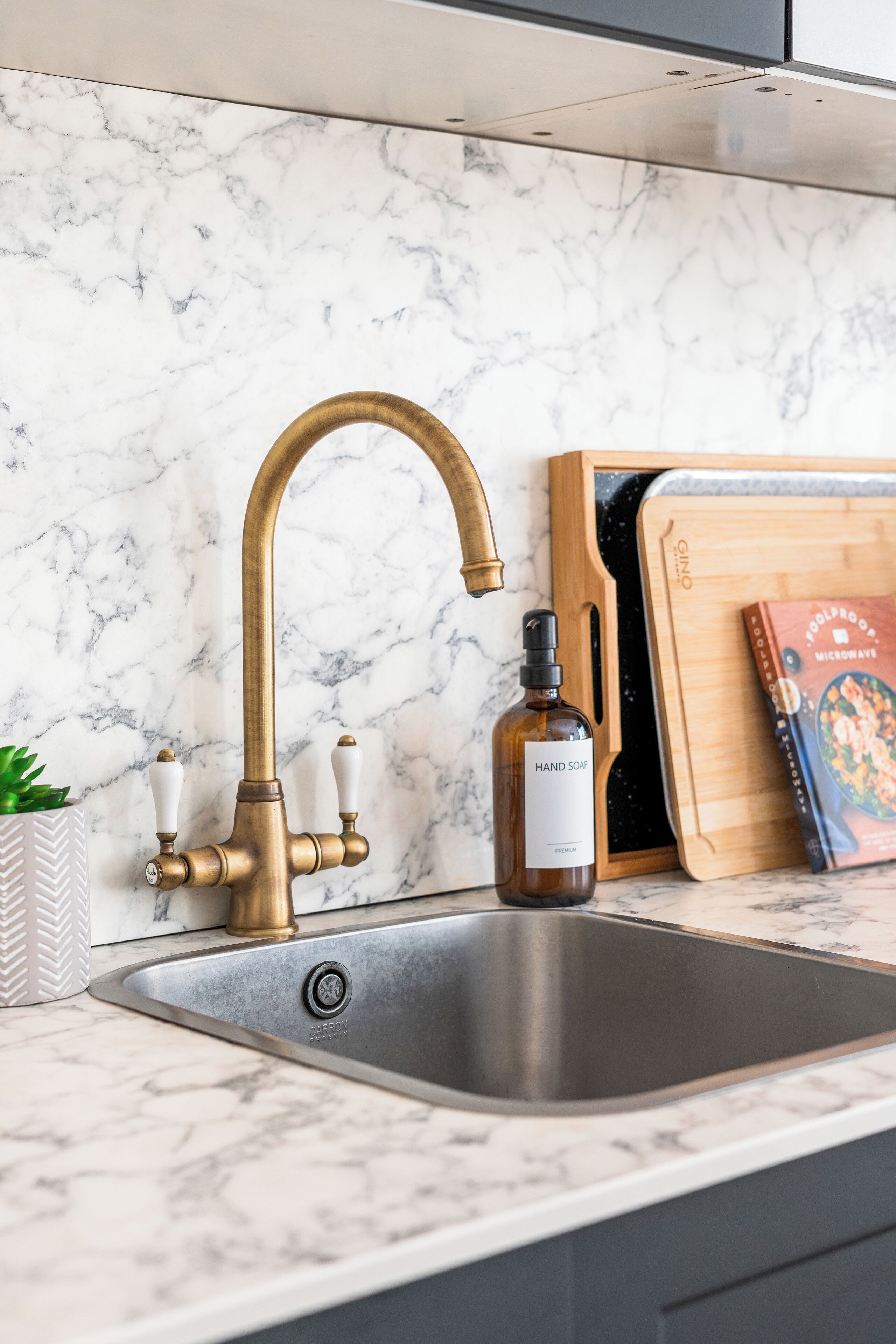 A kitchen with a stainless steel sink, a brass faucet, a wooden cutting board, and a book titled "GOLDPROOF MICOSCARES".