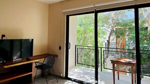 Een moderne kamer met een flatscreen televisie, een houten bureau en een zwarte stoel. Er is een schuifdeur naar een balkon met uitzicht op bomen en een hek.