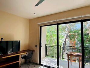 Ein modernes Zimmer mit Flachbildfernseher, einem Holztisch und einem schwarzen Stuhl. Es verfügt über eine Glastür mit Schiebeelement, die auf einen Balkon mit Blick auf Bäume und einen Zaun führt.