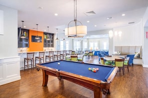 Una mesa de billar en una sala bien iluminada con zona de bar, asientos y vistas al exterior.