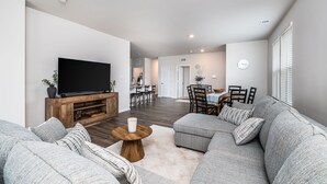 Ett modernt vardagsrum med en stor platt-TV, grå soffor, ett träbord och en matplats i bakgrunden.