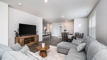 Ein modernes Wohnzimmer mit einem großen Flachbildfernseher, grauen Sofas, einem Holztisch und einem Essbereich im Hintergrund.