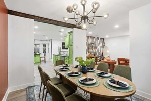 A dining area with a wooden table set for six, surrounded by green upholstered chairs. A chandelier hangs above, and a green kitchen is visible in the background. - The Lone Star Luxe - Pool - Hot Tub - Sport Court (San Antonio)