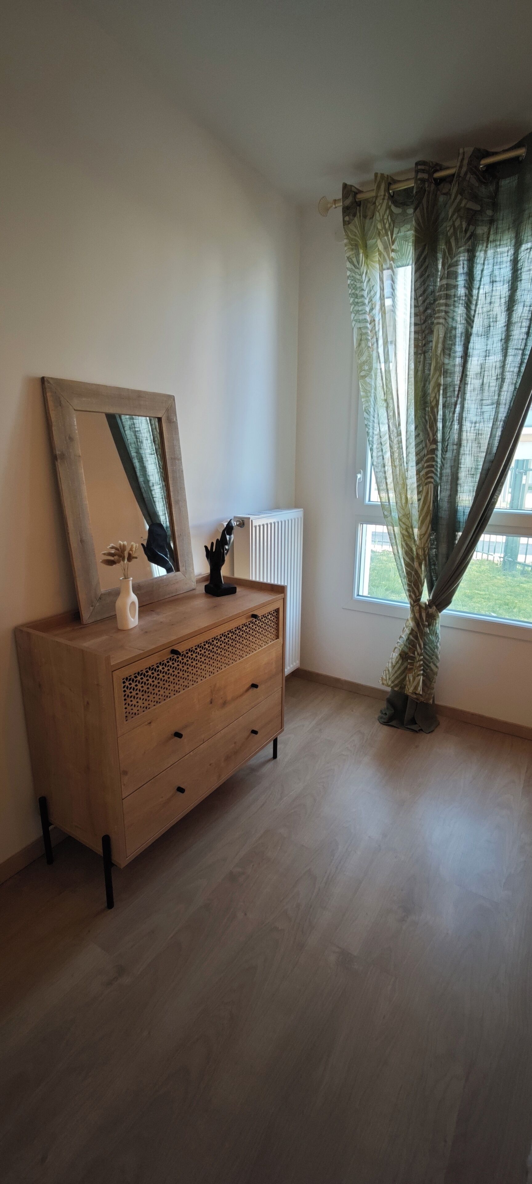 Een kamer met een houten dressoir, een spiegel, een vaas met bloemen en een raam met gordijnen.