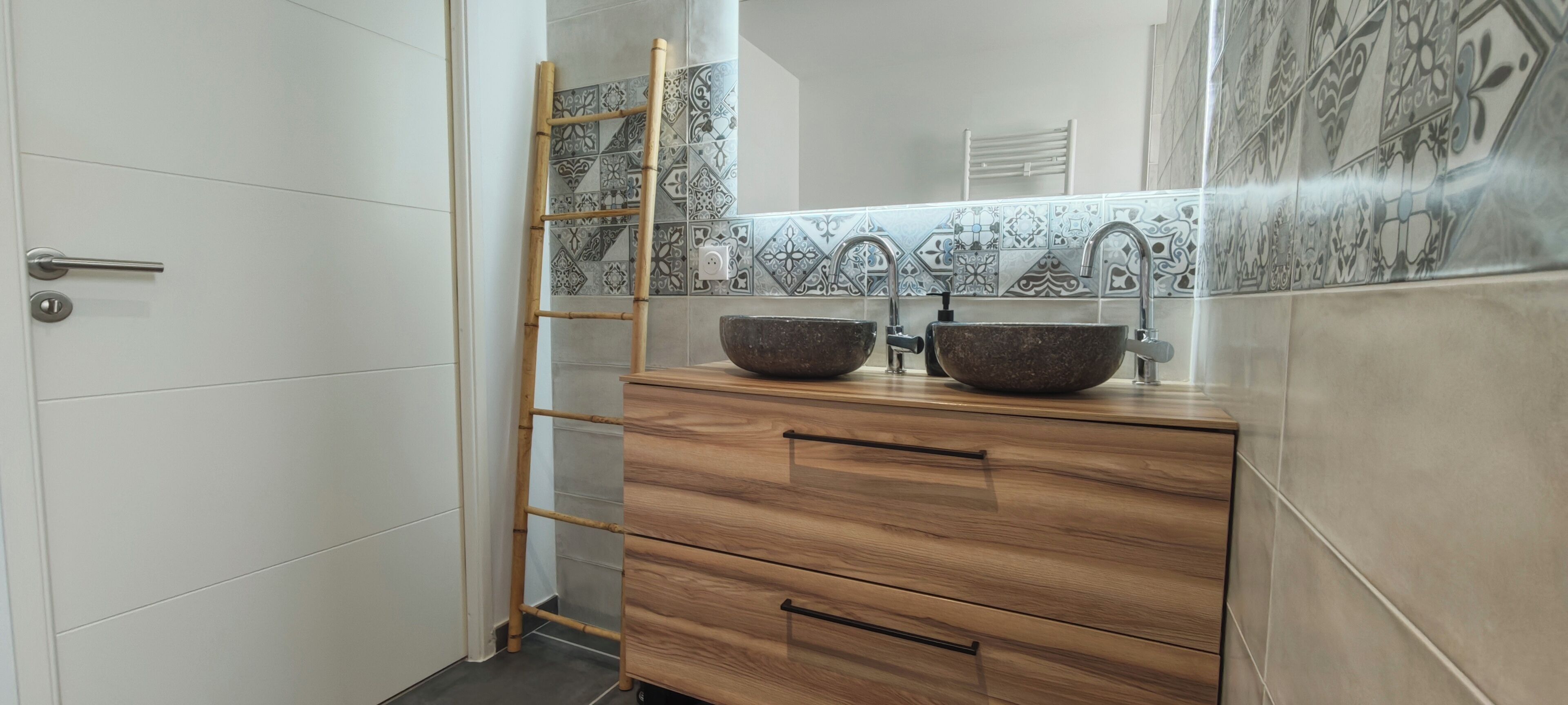 Ein Badezimmer mit zwei Waschbecken, einem Holzkosmetikschrank, einer Bambusleiter und gemusterten Fliesen.