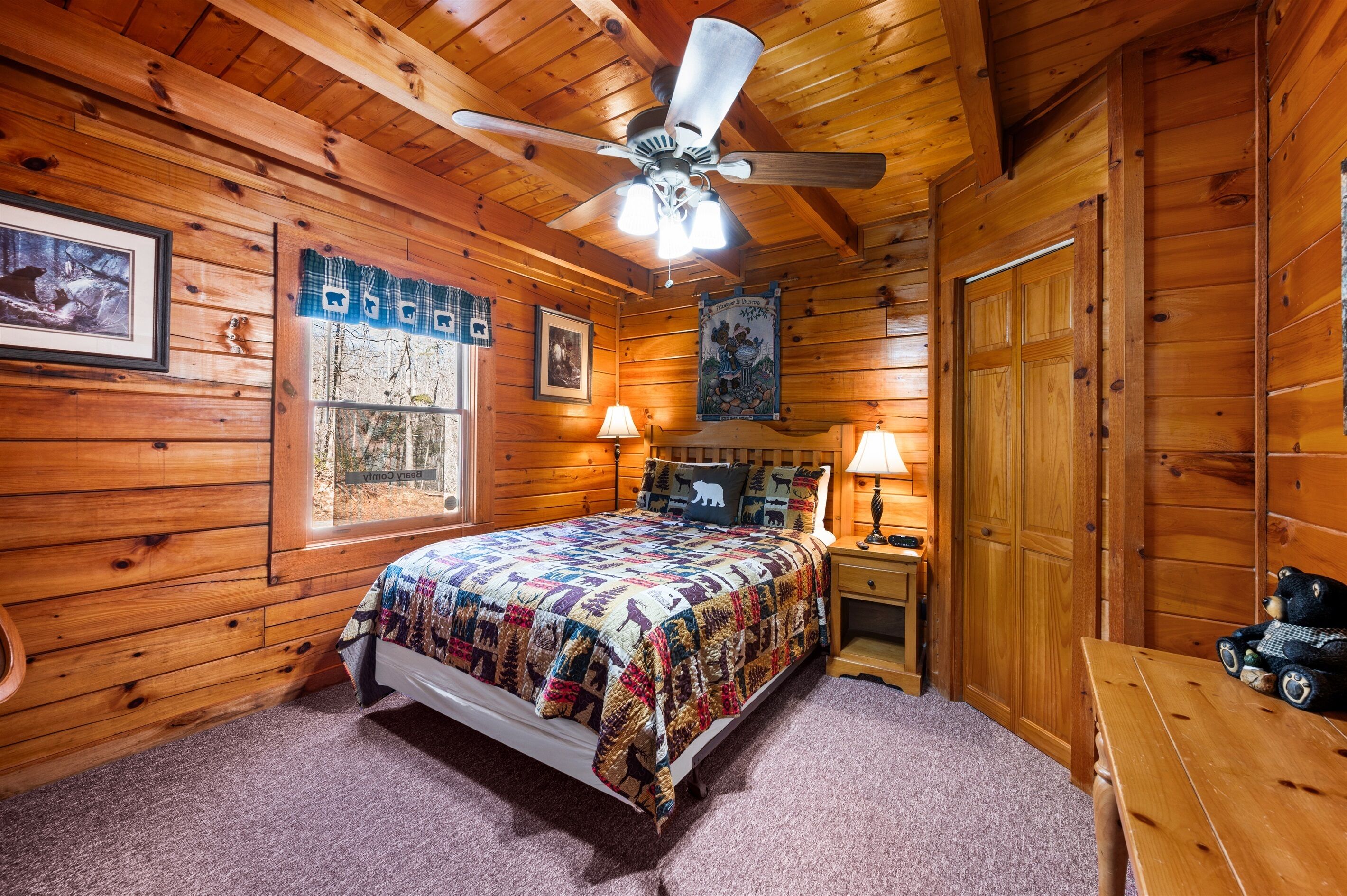 A wooden cabin bedroom with a bed, two bedside lamps, a ceiling fan, and a wooden table with a teddy bear.
