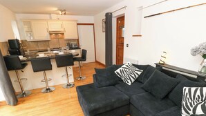 A modern living room with a dark grey sectional sofa, two black bar stools, and a kitchen area with white cabinets and a wooden floor. - Red Kite Cottage (Haverfordwest)