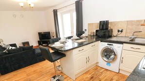 A compact kitchen with a washing machine, a coffee maker, and a dining area with a sofa and chairs. - Red Kite Cottage (Haverfordwest)