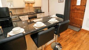 A modern kitchen with white cabinets, a black countertop, and a wooden dining area with bar stools. - Red Kite Cottage (Haverfordwest)