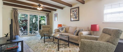 A spacious living room with a sofa, armchairs, and a coffee table. There is a ceiling fan, a TV, and a window with blinds.