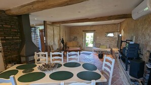 A rustic dining area with a brick floor, wooden beams, and a stone wall. There is a round table with white chairs and a green placemat. A sofa and a small table with a lamp are visible in the background. - Charming country house with garden - 7 guests (Courtemaux)
