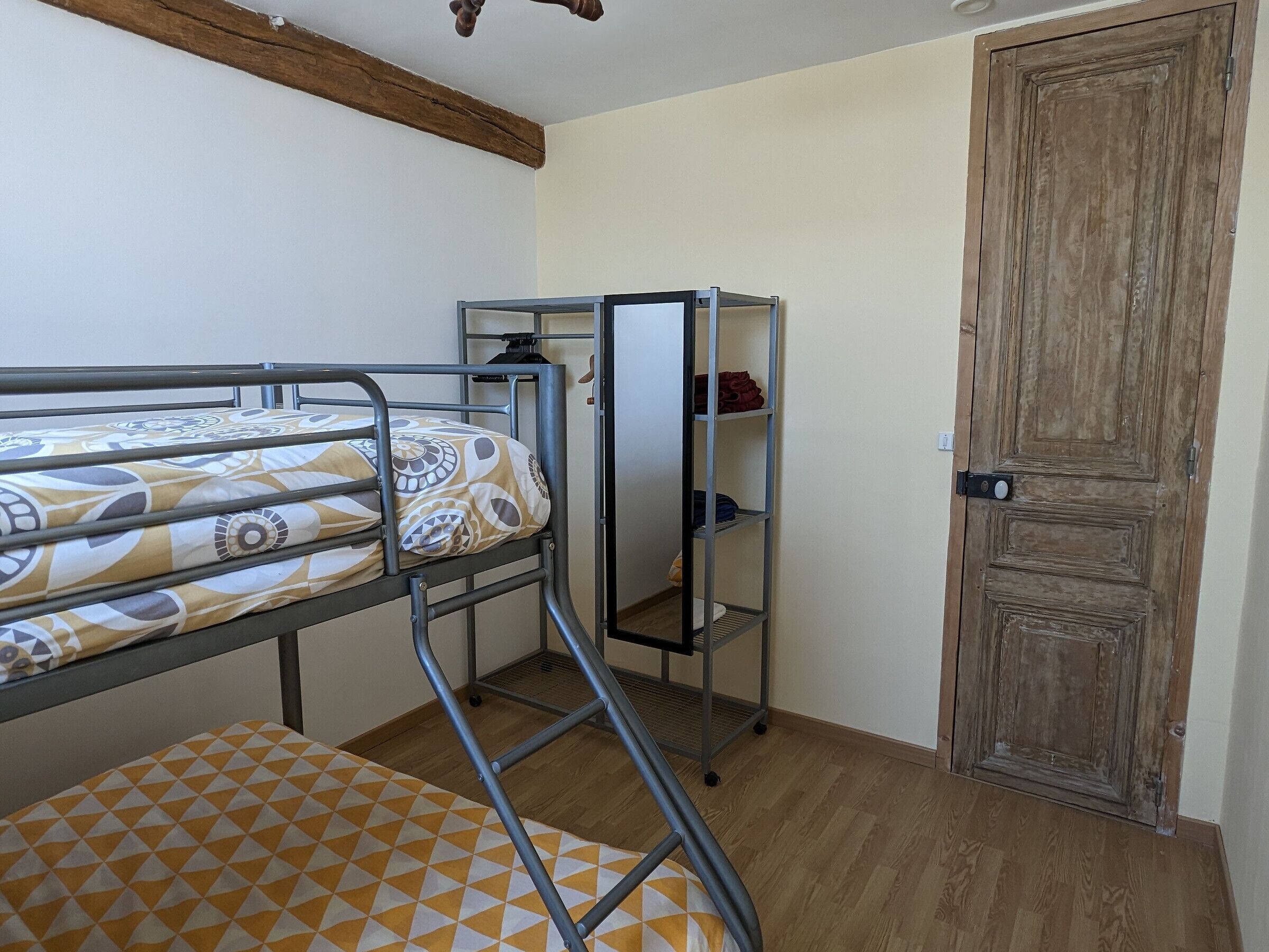 Una cama individual superpuesta con un colchón estampado, una estantería metálica con ropa doblada y una puerta de madera.