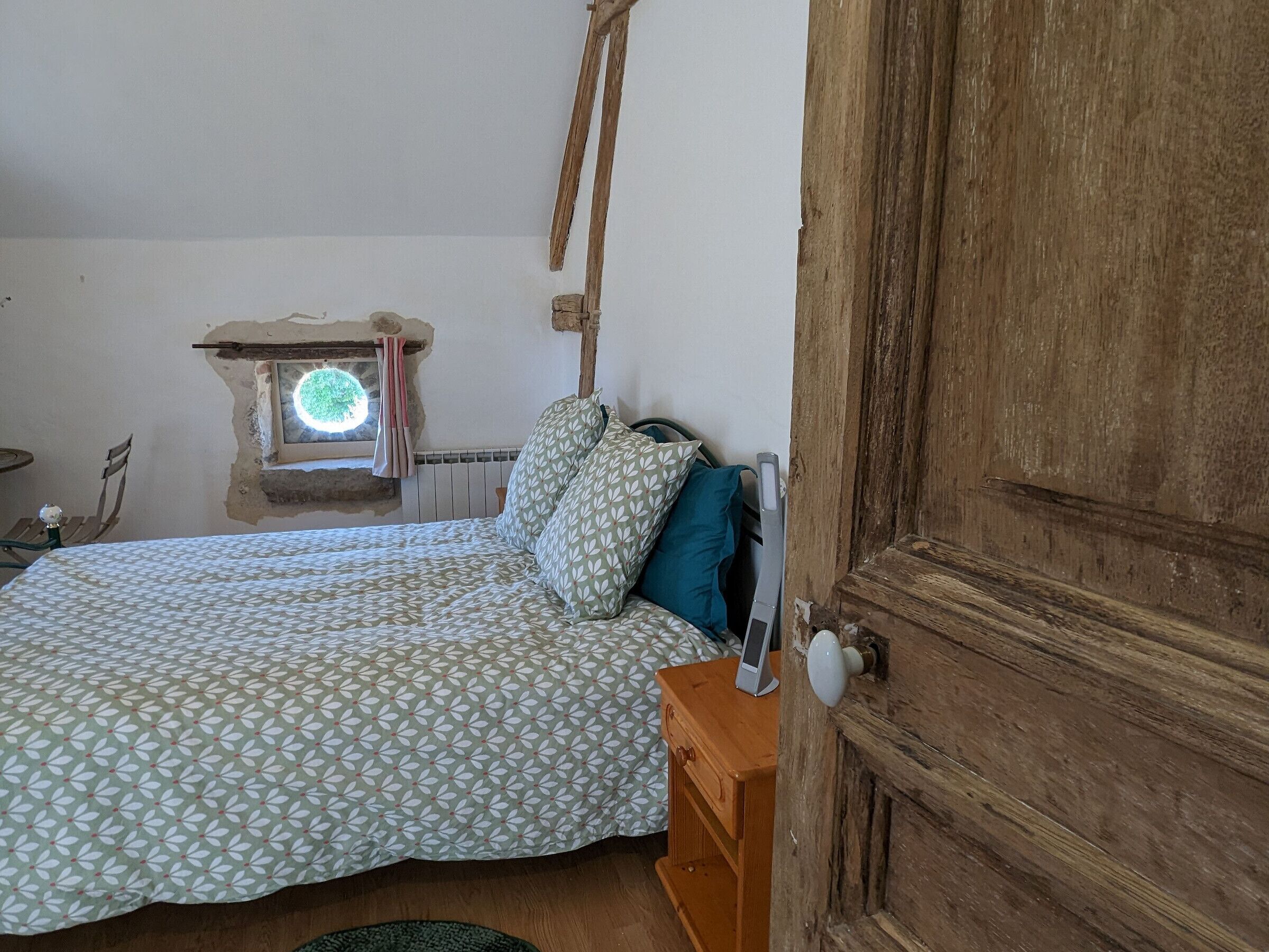 Une chambre à coucher avec un lit, une table de chevet en bois, une porte en bois et une petite fenêtre donnant sur un paysage verdoyant.