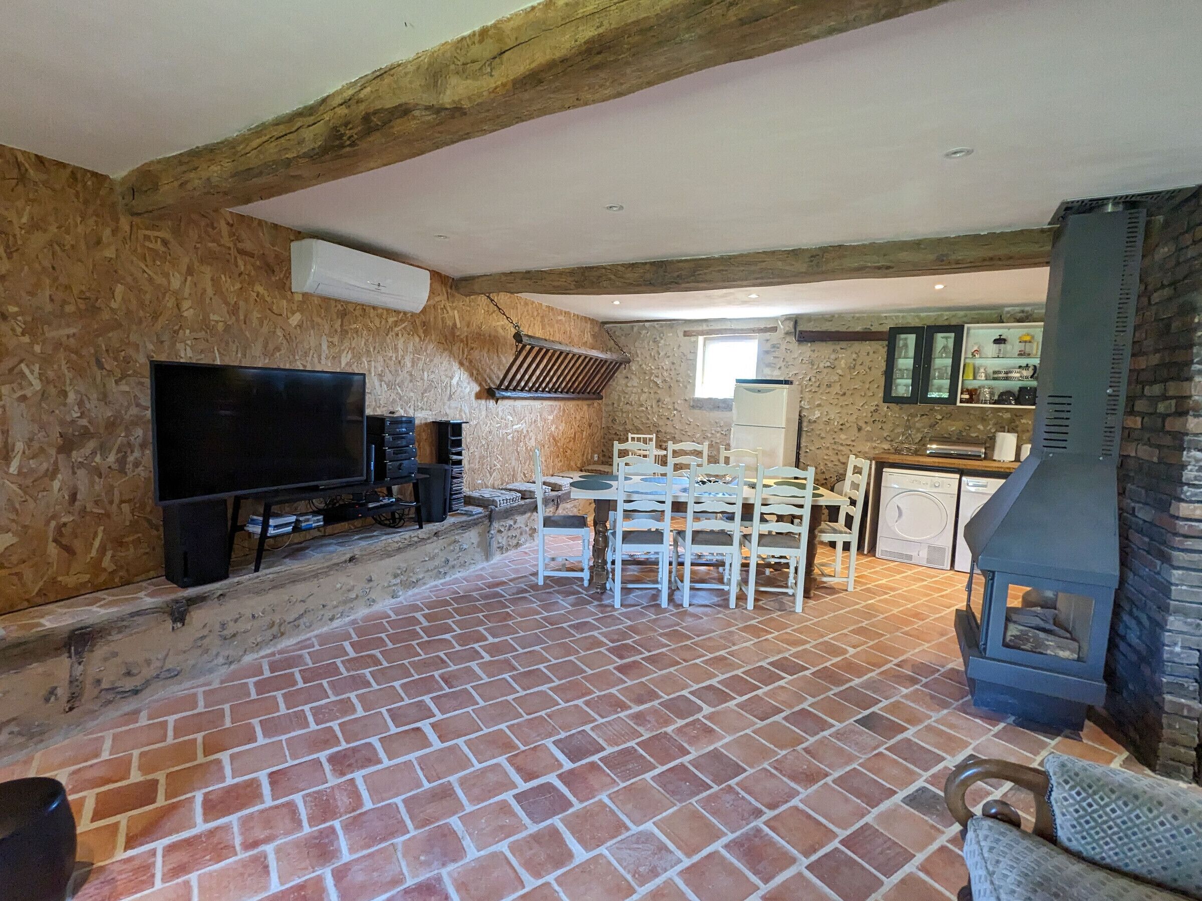 Una habitación rústica con vigas de madera a la vista, suelo de ladrillo, televisión, una mesa de comedor con sillas y una zona de cocina con una parrilla.