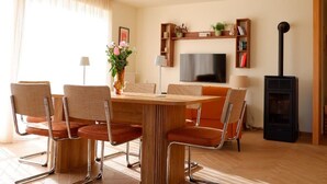 A modern living room with a wooden dining table, orange chairs, a flat-screen TV, a black wood-burning stove, and a bookshelf. - Mid-Century Stay in Val-d'Ajol with Parking and Pets Welcome (Le Val-d'Ajol)