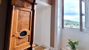 Un reloj de pie de madera con esfera blanca y números romanos, situado junto a una ventana con vistas al paisaje.
