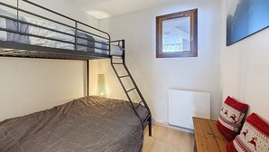 En våningssäng med en stege, ett litet bord, ett element och ett fönster med utsikt över en byggnad.