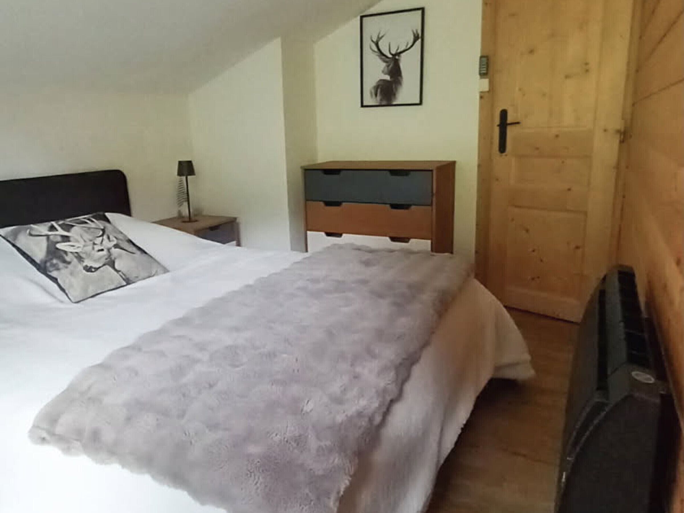 A bedroom with a bed, a nightstand, a lamp, a wooden headboard, a framed deer picture, and a wooden door.