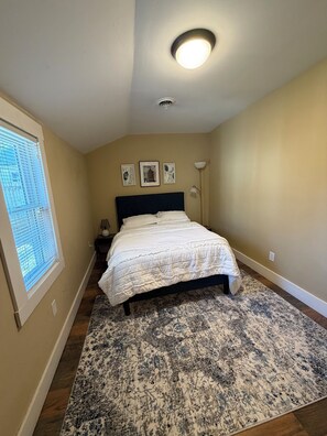 Un dormitorio con cama, una ventana con persianas, una lámpara de pared y tres cuadros enmarcados.