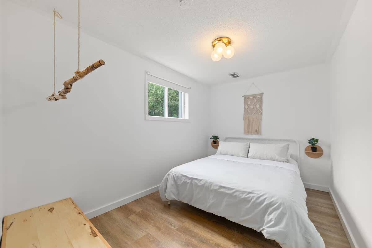A bedroom with a bed, a window, a hanging light fixture, and a small wooden table.