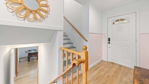 Una escalera con barandillas de madera que lleva a una plataforma, una puerta blanca con un ventanal y un espejo decorativo fijado a la pared.