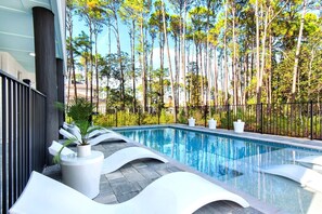 Una piscina con sillas de descanso blancas y macetas, rodeada de árboles altos y una valla negra.
