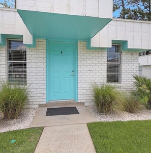 Un bâtiment avec une porte turquoise, des murs en briques blanches et un petit jardin devant, avec de l’herbe et des plantes.