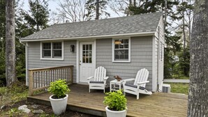 A small cabin with a porch, two Adirondack chairs, and potted plants. - Amore | Romantic Coastal Cottage Near Spruce Point (Boothbay Harbor)