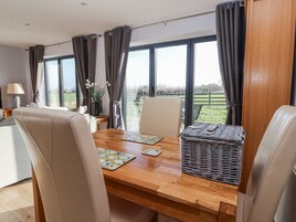 A dining area with a wooden table, two chairs, a woven basket, and a view of a green landscape through large windows.