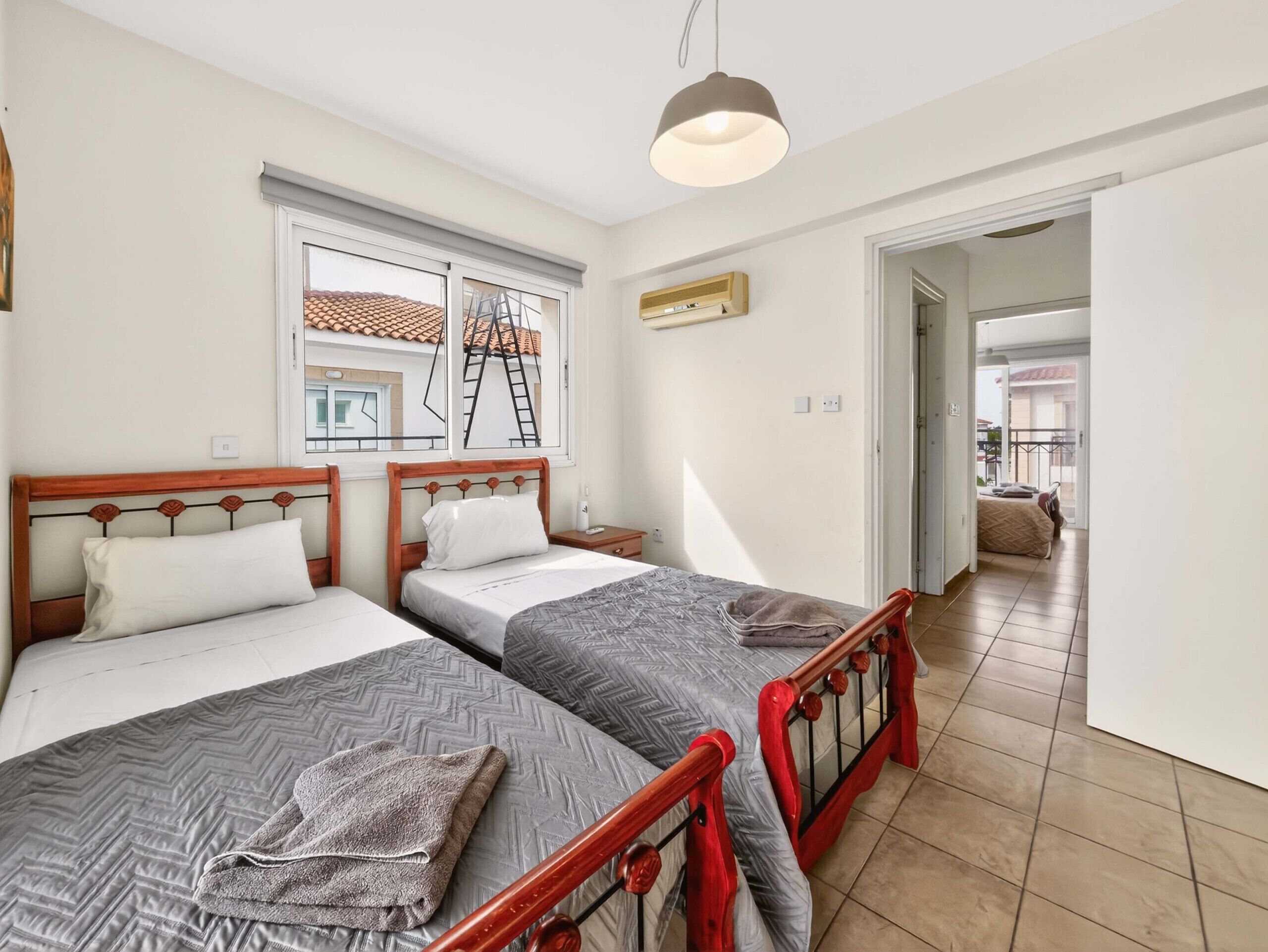 Un dormitorio con dos camas individuales, una ventana con vista a un balcón, una lámpara en el techo y una puerta que da a otra habitación.