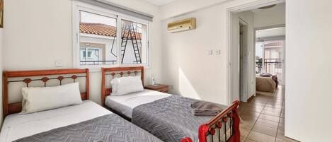 Un dormitorio con dos camas individuales, una ventana con vista a un balcón, una lámpara en el techo y una puerta que da a otra habitación.