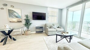 A modern living room with a flat-screen TV, a sofa, a coffee table, and a dining area with a glass table and chairs. -  The Coastal - Gulf View 8th floor condo at Bonita Springs Beach & Tennis Club (Bonita Springs)