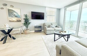 A modern living room with a flat-screen TV, a sofa, a coffee table, and a dining area with a glass table and chairs. -  The Coastal - Gulf View 8th floor condo at Bonita Springs Beach & Tennis Club (Bonita Springs)