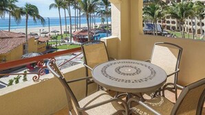 A balcony with a round table and chairs, overlooking a beach with palm trees and a resort building. - Cozy 1-bedroom deluxe condo with AC in lovely San José del Cabo (San José del Cabo)