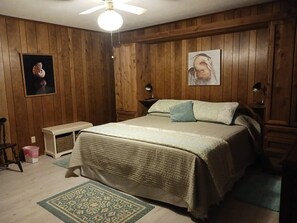 A bedroom with a bed, a nightstand, a lamp, a small table, and a wall-mounted shelf.