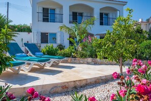 Um edifício de dois andares com varanda, área de piscina com espreguiçadeiras e jardim com diversas plantas e flores.