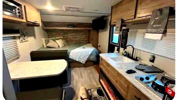 Interior compacto de um veículo recreativo, com cozinha compacta, cama e pequena área de refeições.