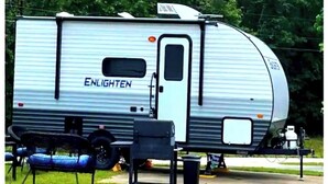 Uma caravana prateada com a marca 'ENLIGHTEN' na lateral, equipada com uma pequena cozinha acoplada e um sanitário portátil.