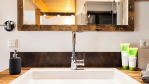 Casa de banho com lavatório branco, torneira cromada e espelho com moldura de madeira. Existem duas embalagens de higiene pessoal verdes no balcão.